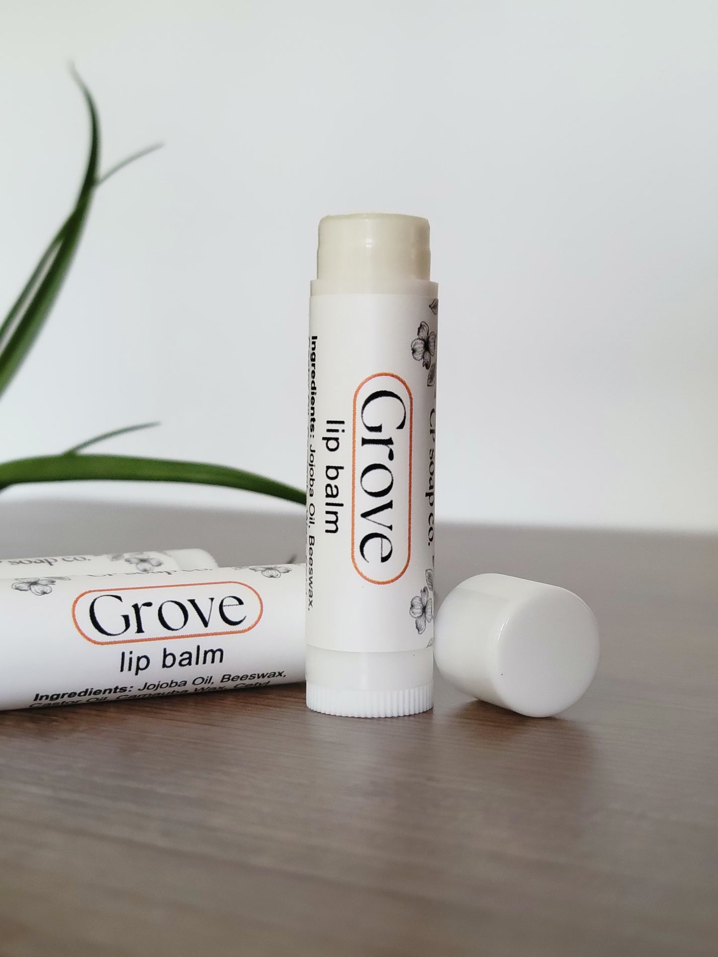 Two Grove lip balm tubes on a light wooden surface with a plant in the background.