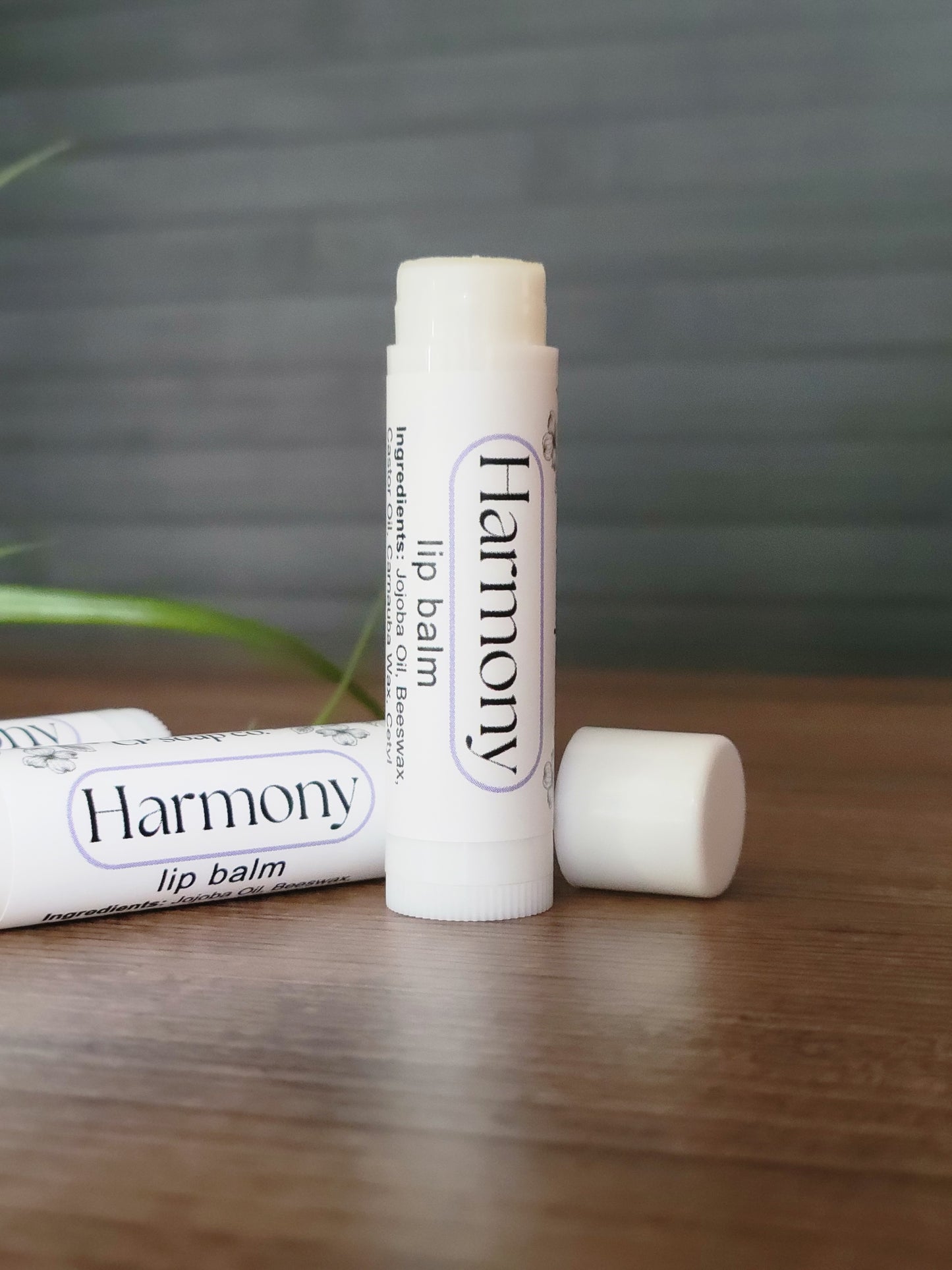 Two lip balms labeled 'Harmony' on a wooden surface with a blurred background.