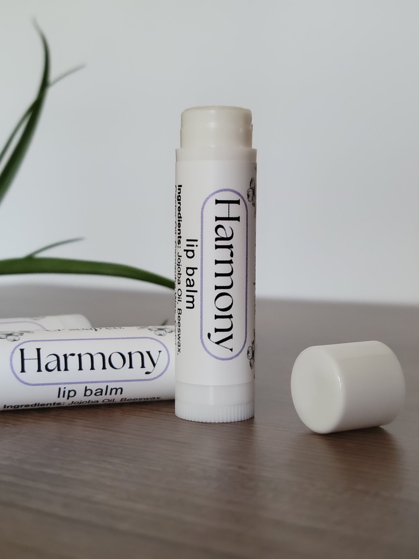 Two lip balms labeled 'Harmony' on a light wooden surface with a plant in the background.