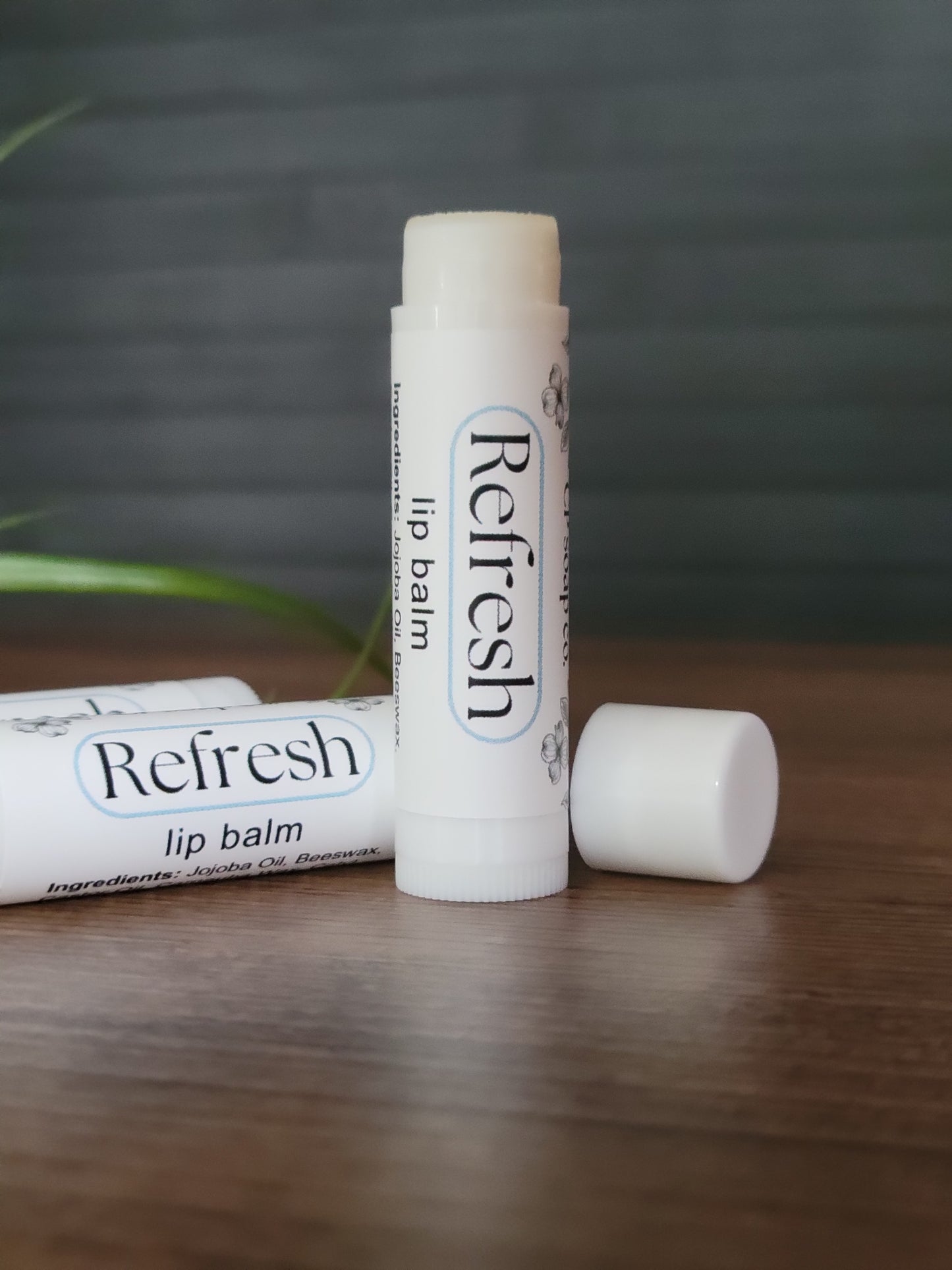 Two lip balms labeled 'Refresh' on a wooden surface with a blurred background.