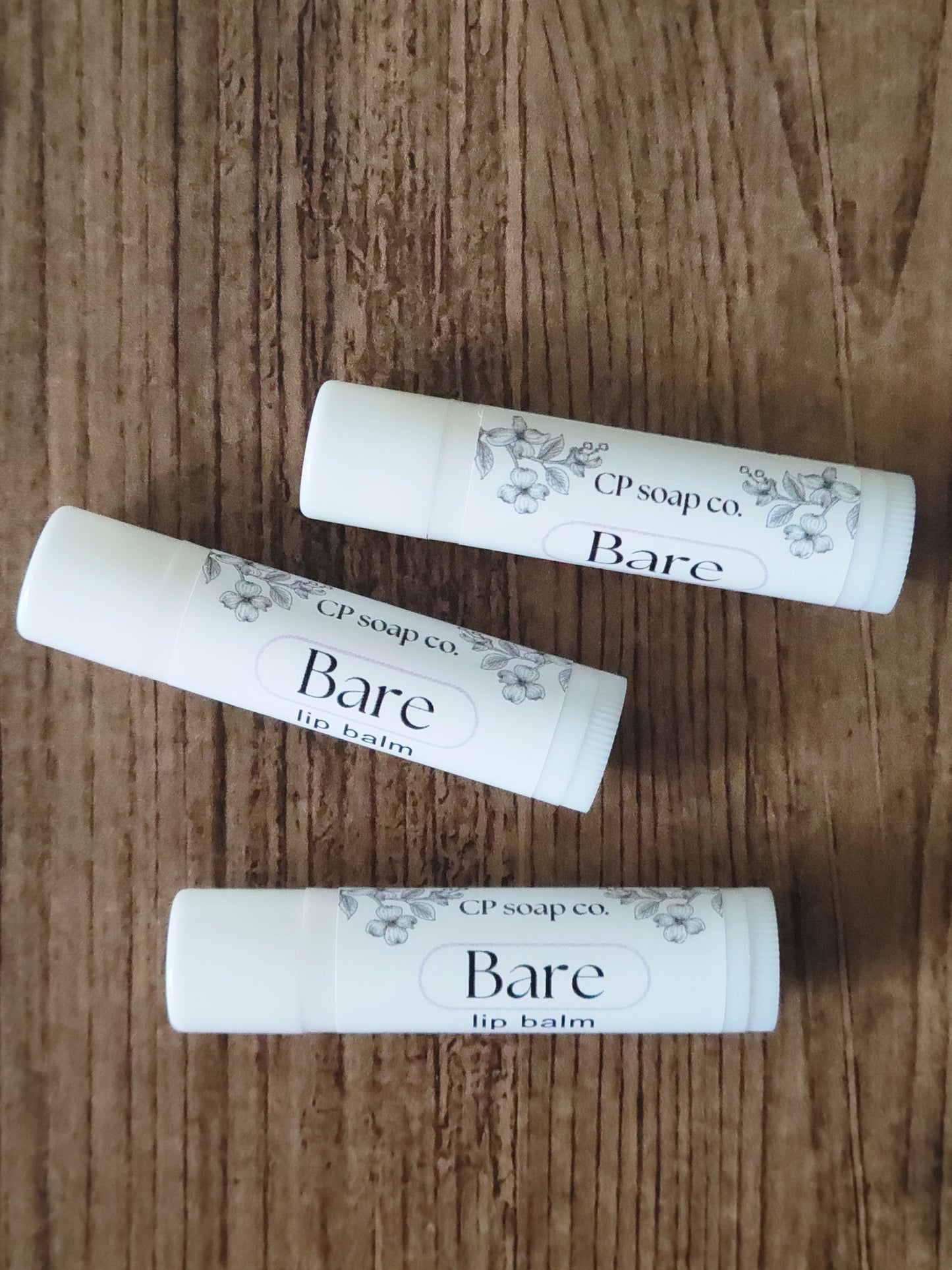 Three lip balm tubes labeled 'Bare' from CP Soap Co on a wooden surface.
