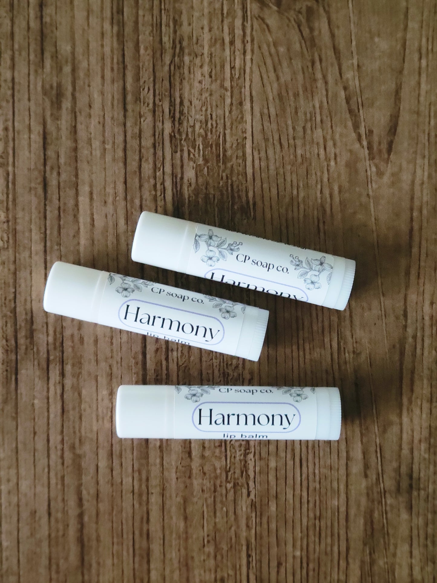Three lip balm tubes labeled 'Harmony' on a wooden surface