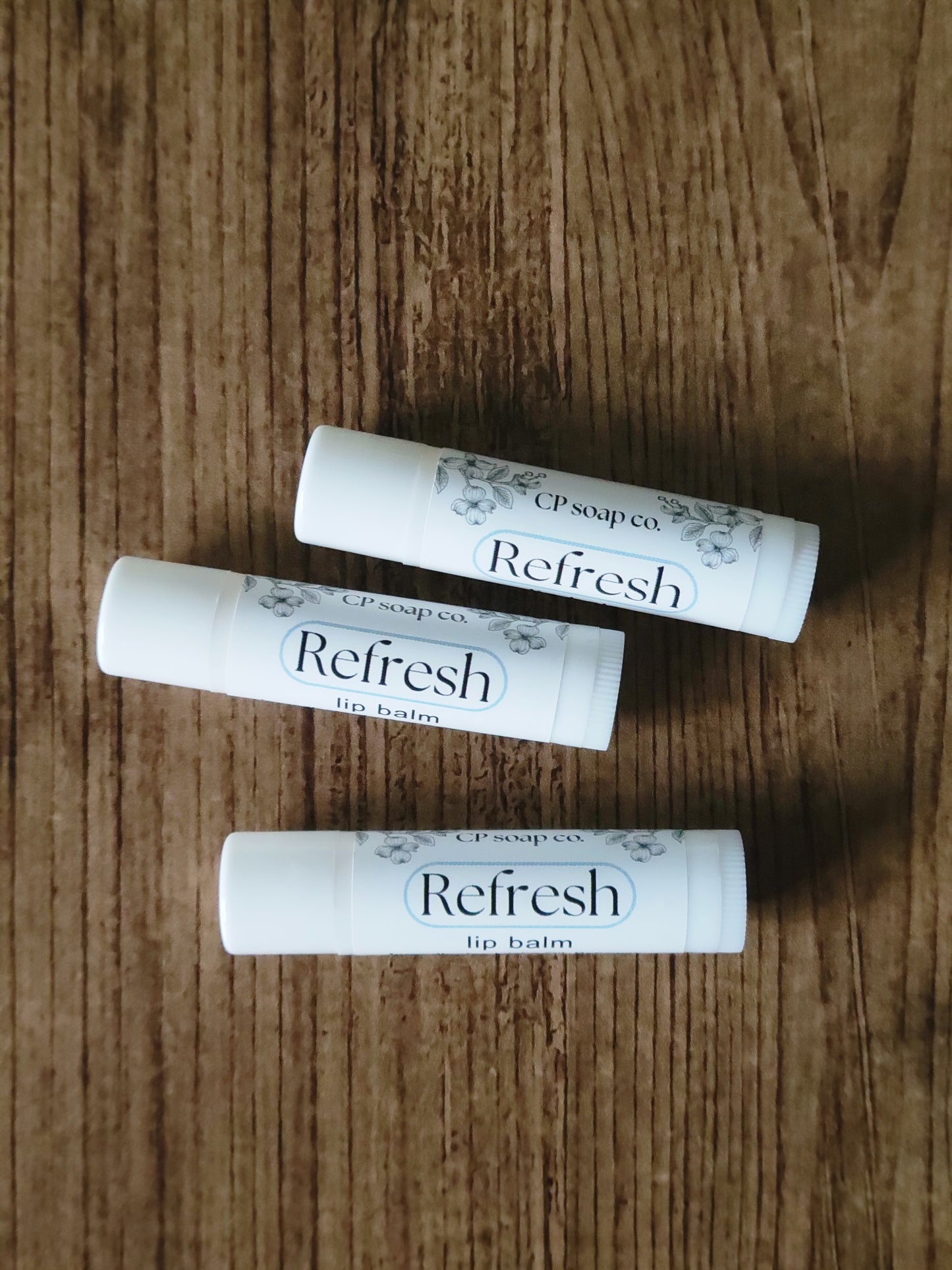 Three lip balm tubes labeled 'Refresh' on a wooden surface