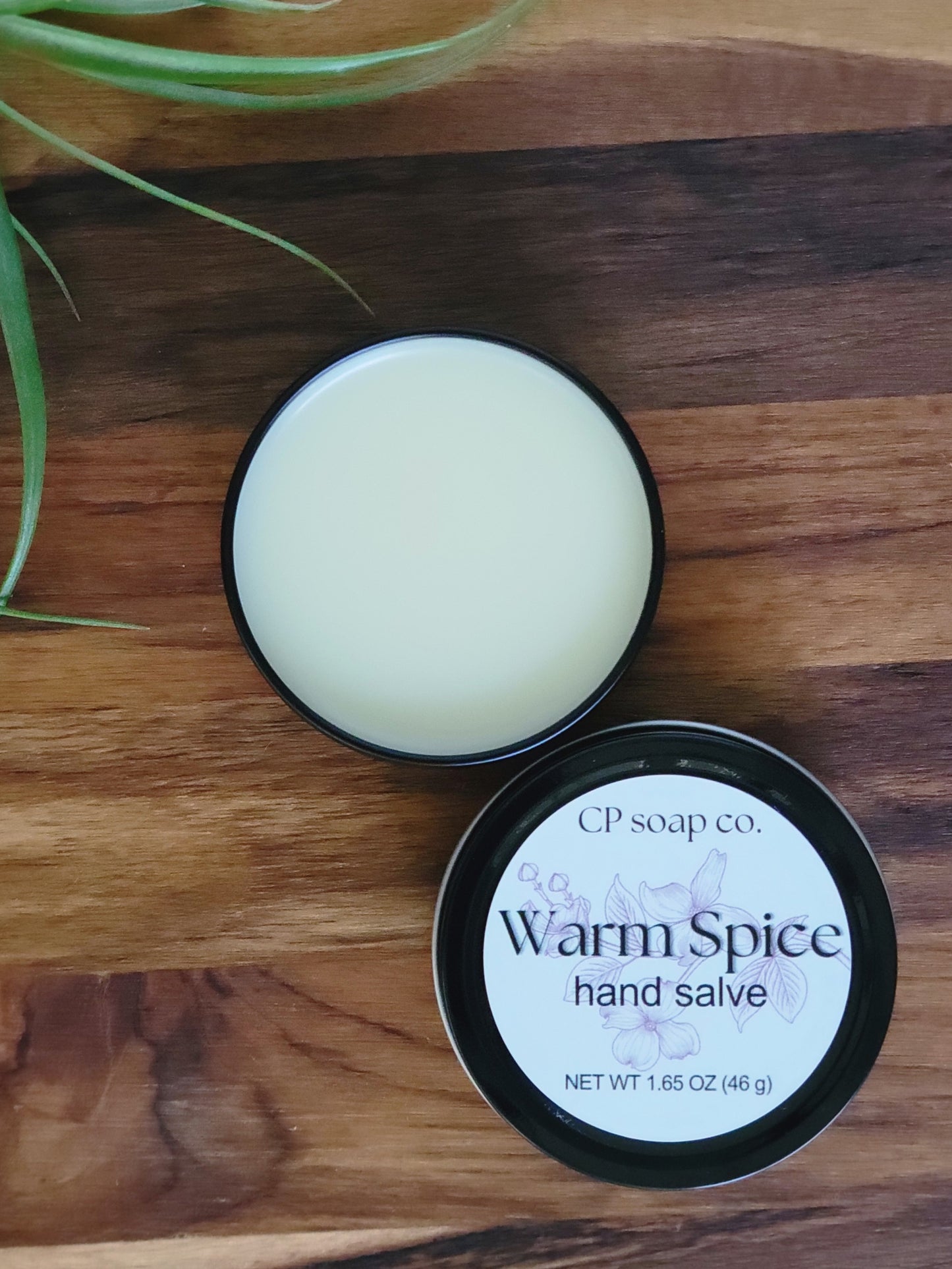 CP Soap Co. Warm Spice hand salve on a wooden surface with a plant in the corner.