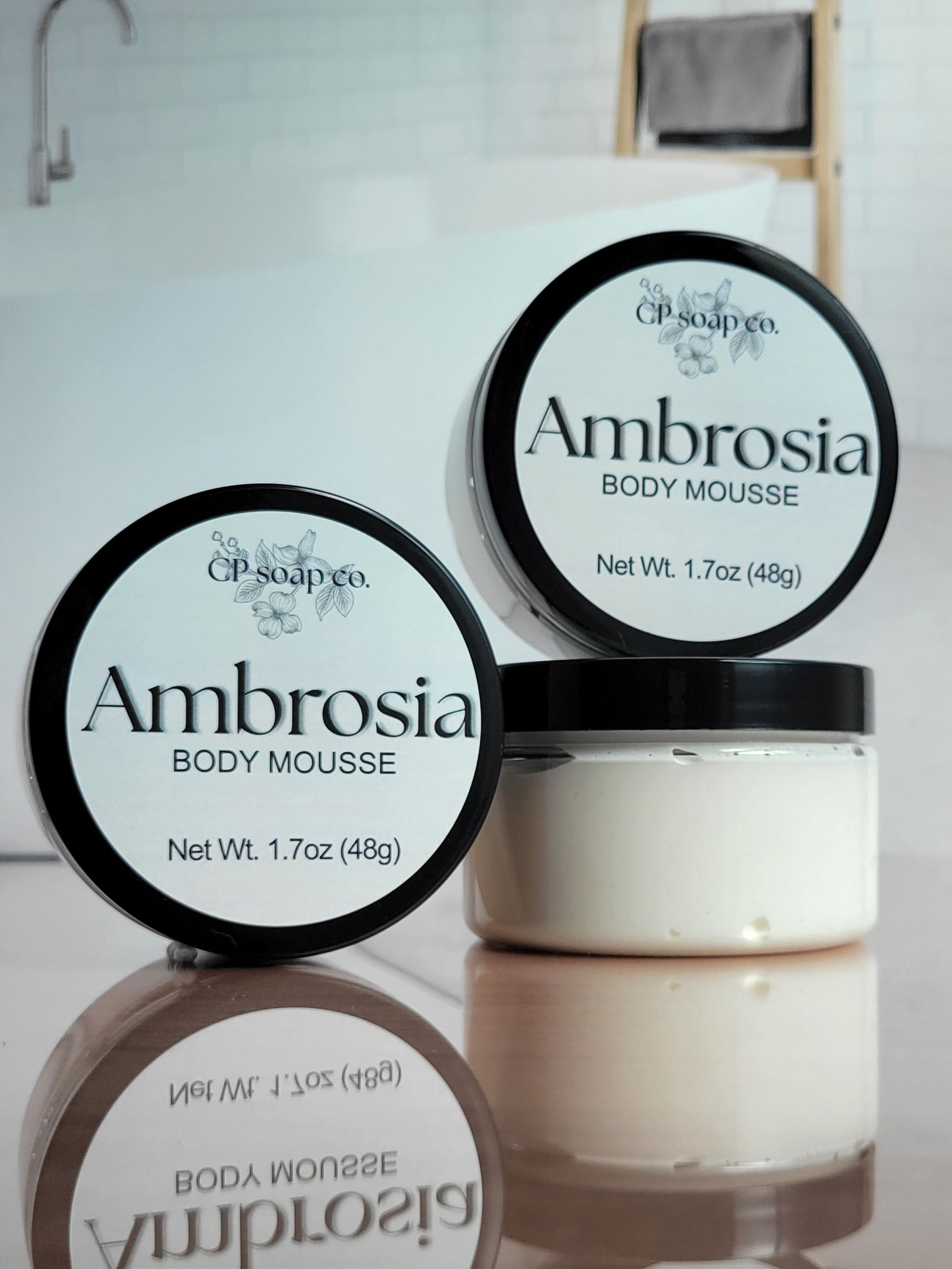 Two containers of Ambrosia body mousse with visible branding on a reflective surface.