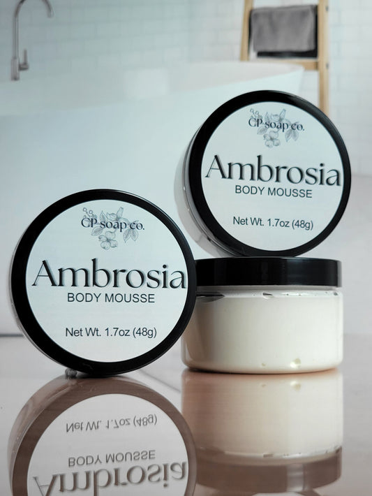 Two containers of Ambrosia body mousse with visible branding on a reflective surface.