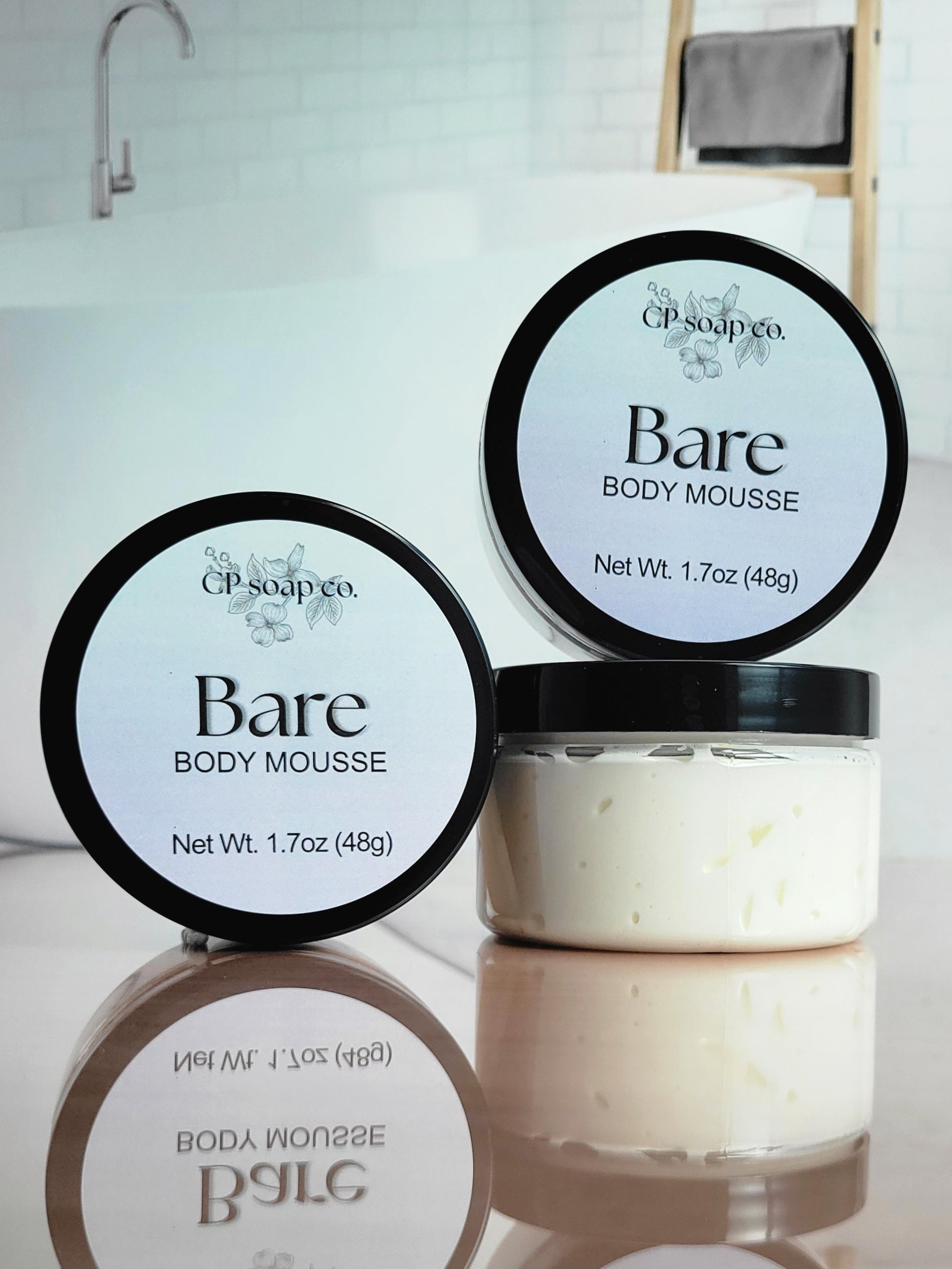 CP Soap Co. Bare Body Mousse containers on a reflective surface with a blurred background.