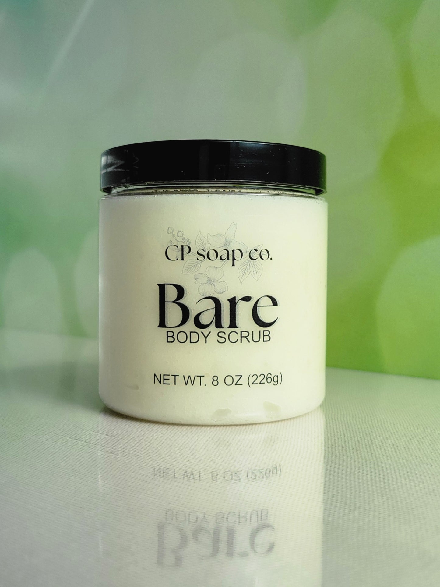 CP Soap Co. Bare Body Scrub jar on a reflective surface with a blurred green background