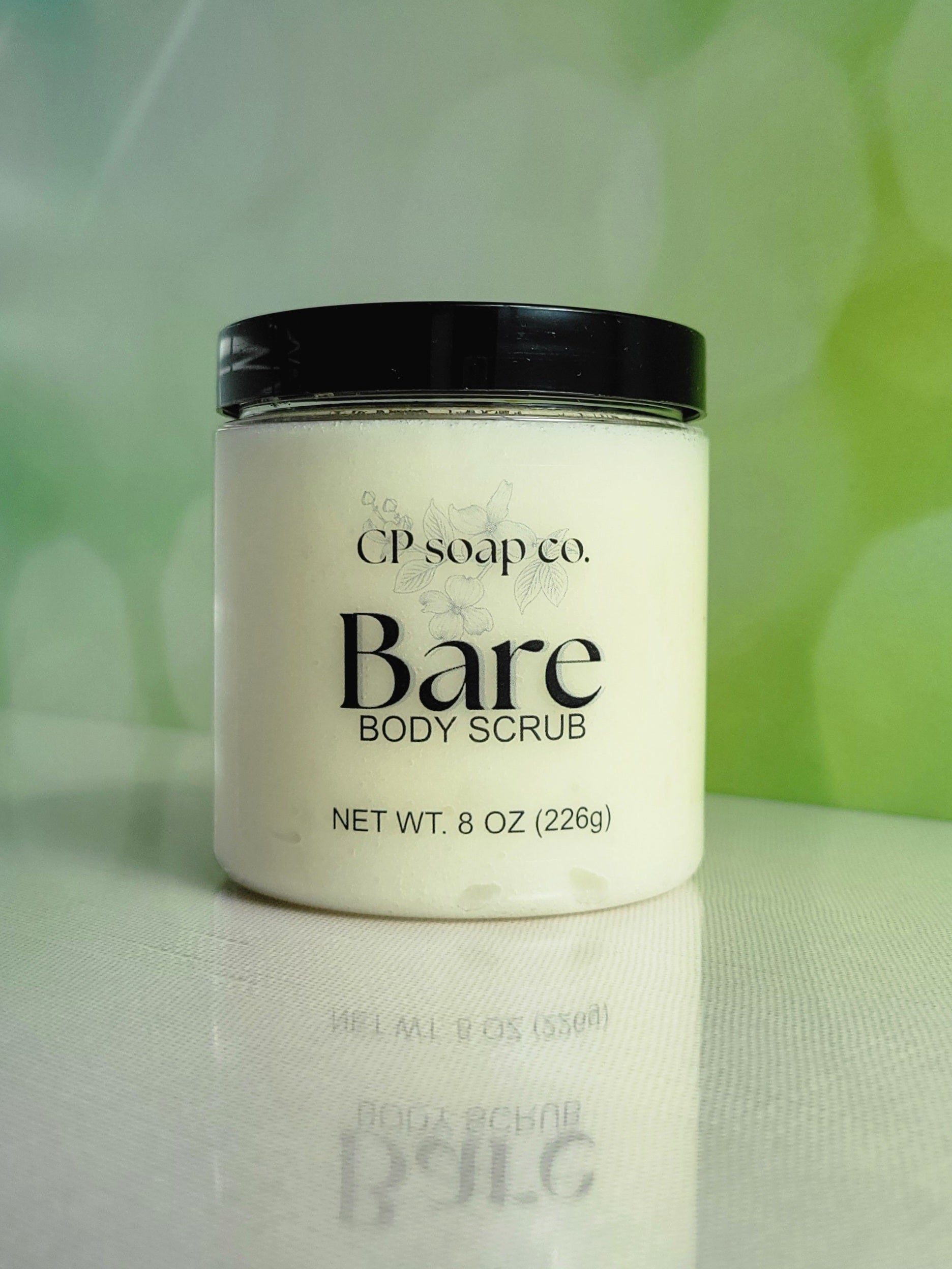 CP Soap Co. Bare Body Scrub jar on a reflective surface with a blurred green background