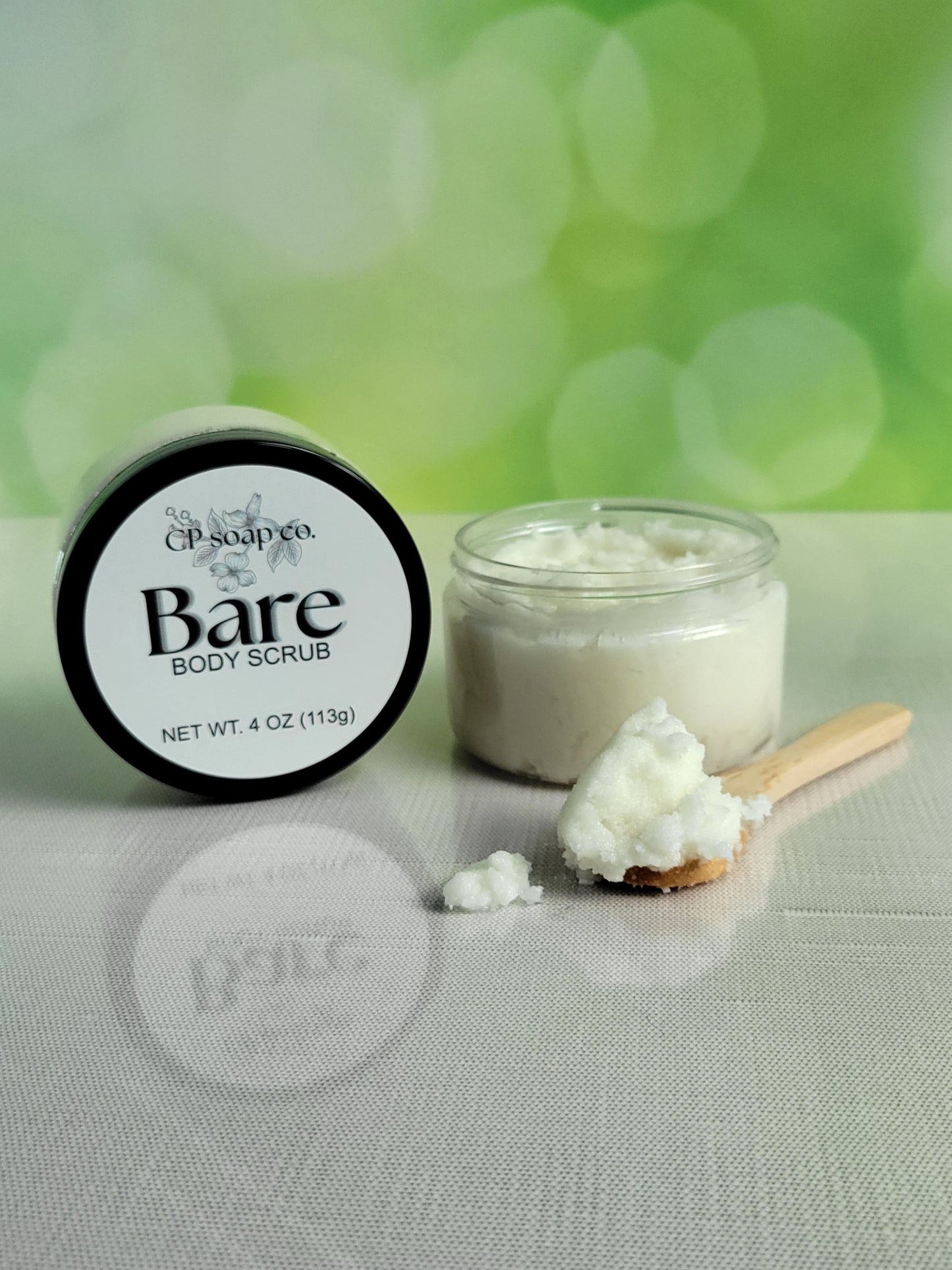 Jar of Bare Body Scrub with a wooden scoop on a light surface with a green blurred background