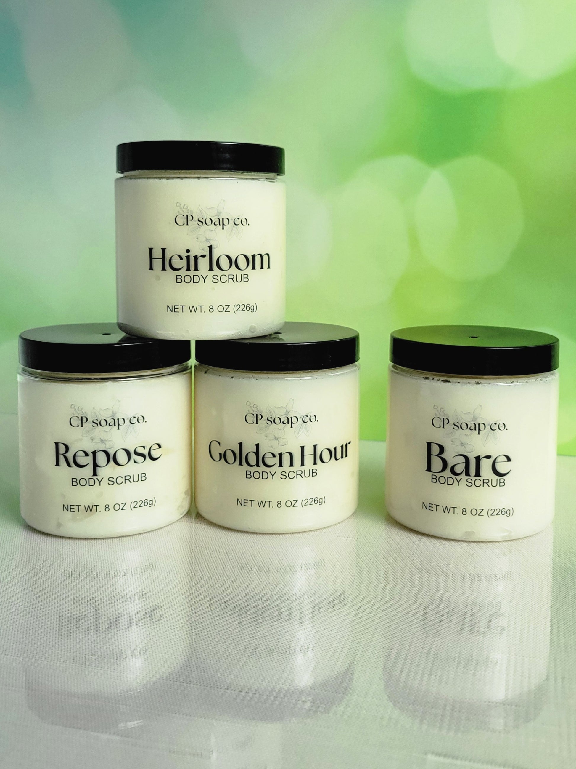 Four jars of CP Soap Co. body scrubs on a reflective surface with a green blurred background