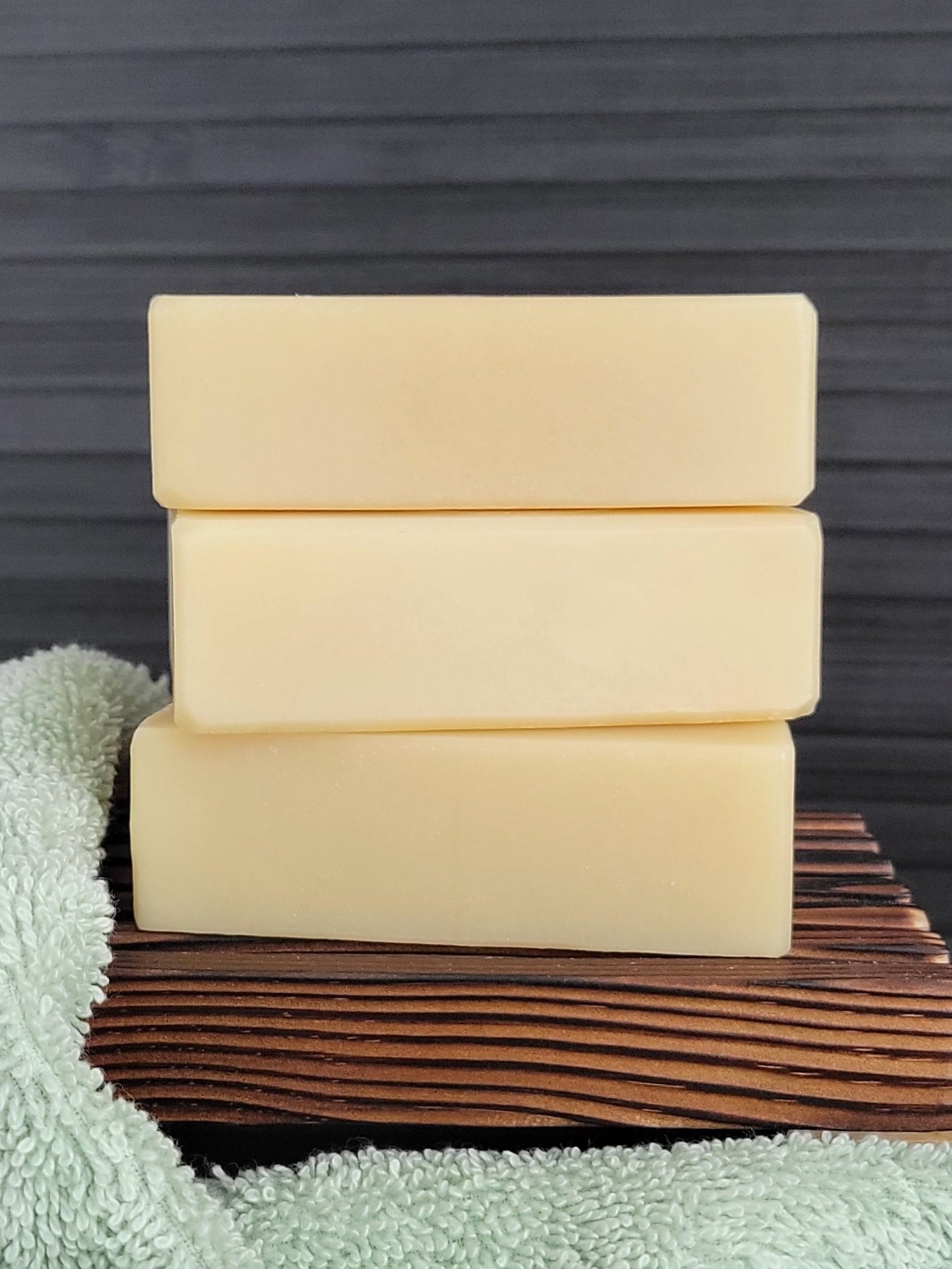 Three bars of soap stacked on a wooden surface with a towel underneath, against a dark background.