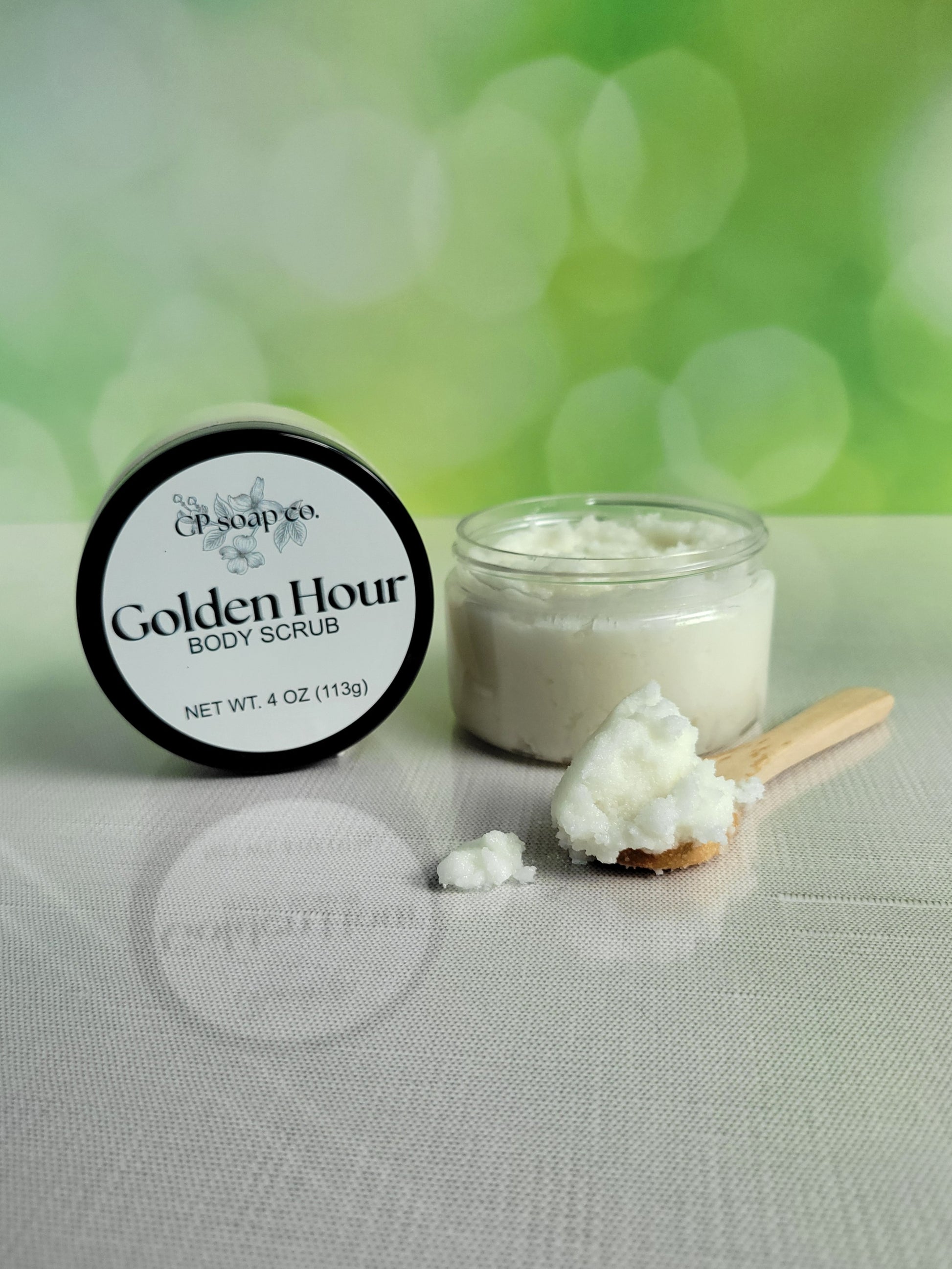 Golden Hour body scrub with a jar and scoop on a light surface with a green blurred background