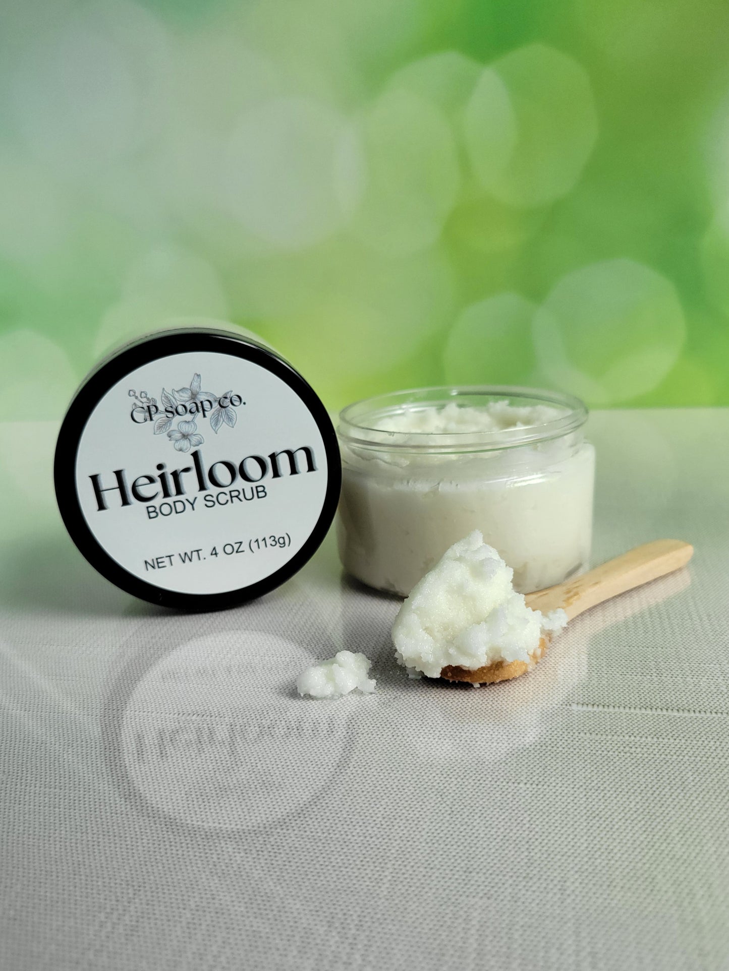 Heirloom body scrub with a jar and scoop on a textured surface with a green blurred background