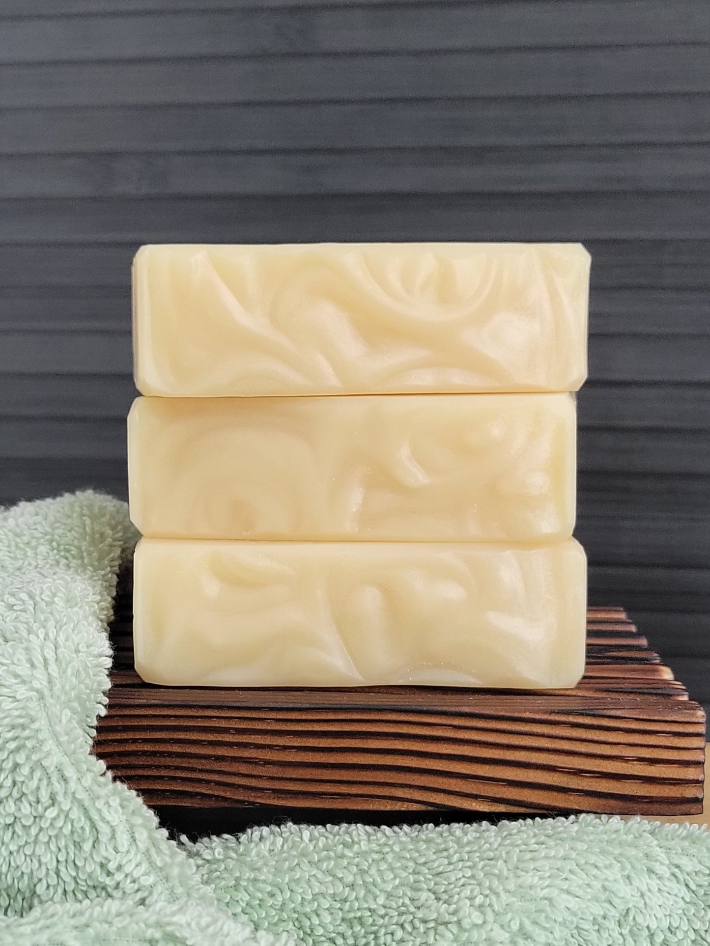 Three bars of soap stacked on a wooden block with a green towel underneath against a dark background